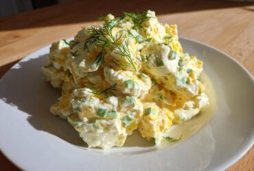 A generous serving of creamy egg salad recipe, mixed with green onions and topped with fresh dill, sits on a white plate.