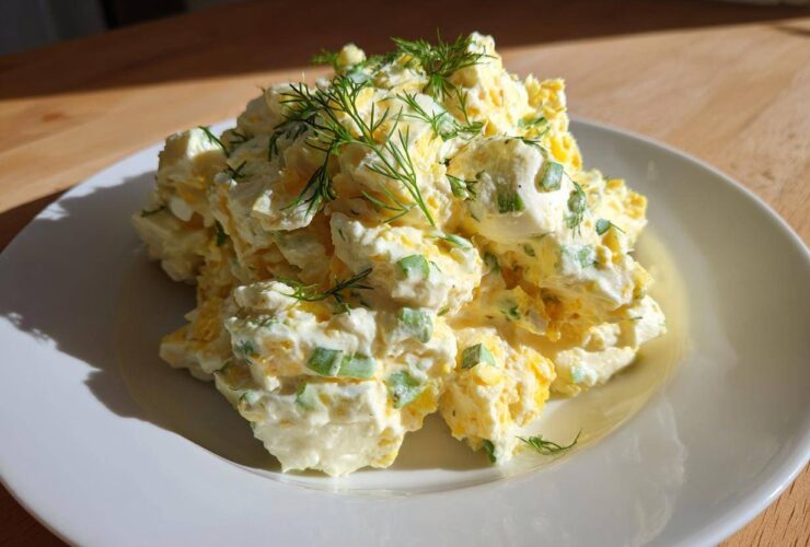 A generous serving of creamy egg salad recipe, mixed with green onions and topped with fresh dill, sits on a white plate.