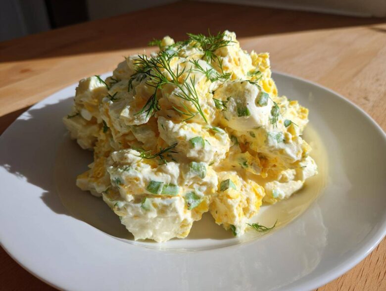 A generous serving of creamy egg salad recipe, mixed with green onions and topped with fresh dill, sits on a white plate.