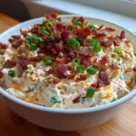 A white bowl filled with creamy Million Dollar Dip, topped generously with crumbled bacon and sliced green onions.