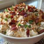 A white bowl filled with creamy Neiman Marcus dip, topped generously with crumbled bacon, sliced almonds, and green onions.