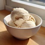 Three scoops of creamy, light brown protein ice cream served in a white bowl near a sunlit window.