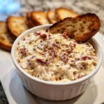 Close-up of a warm, creamy reuben dip topped with melted cheese and spices, served with toasted bread slices.