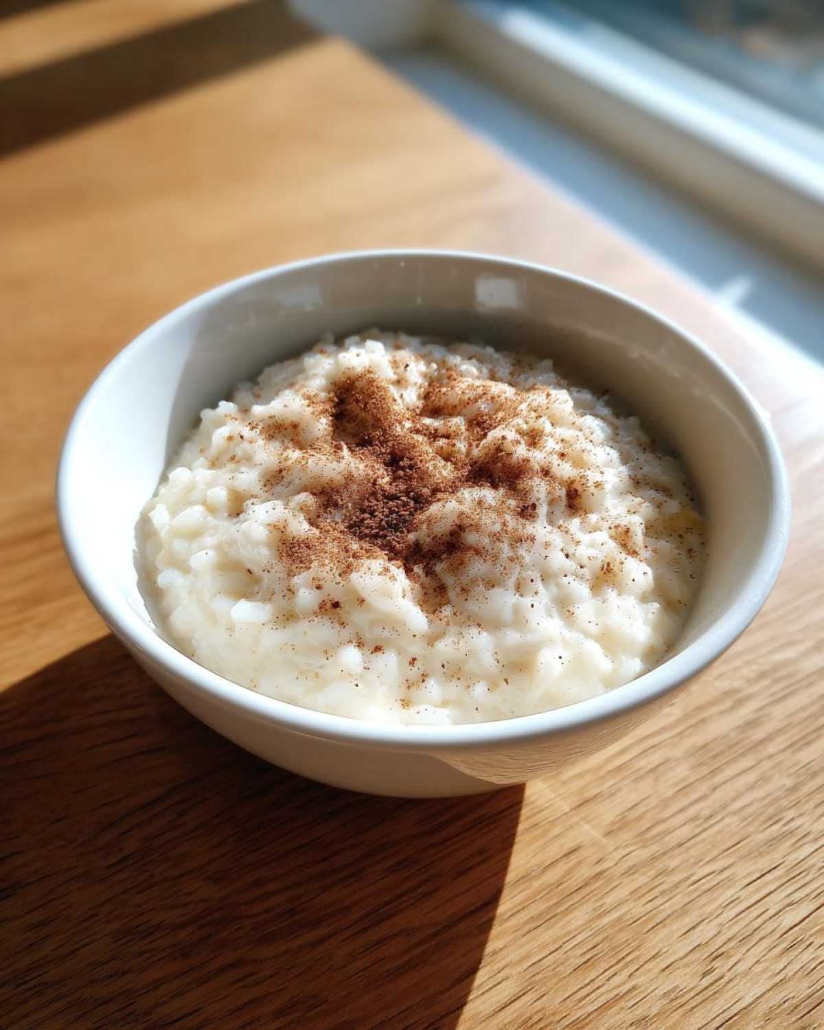 A white bowl filled with creamy rice pudding topped generously with ground cinnamon, sitting on a wooden table near a window.
