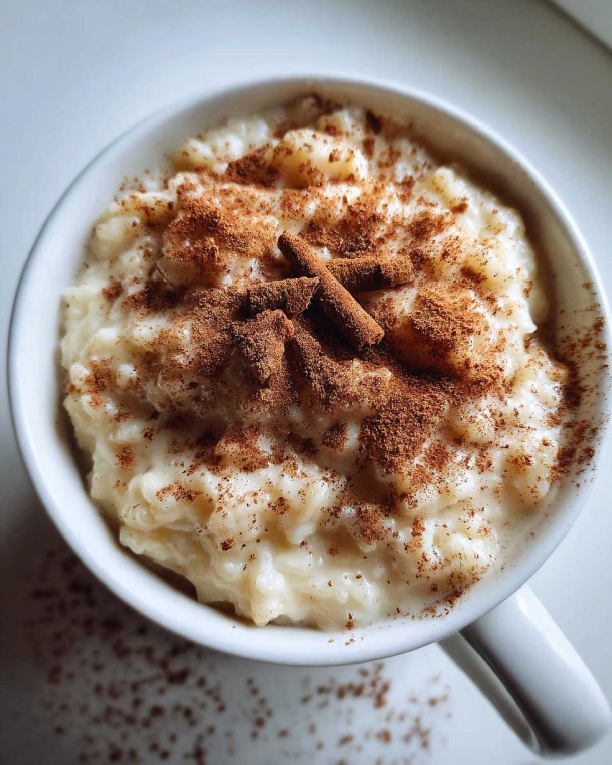 Overhead close-up of creamy rice pudding recipe served in a white mug, generously dusted with ground cinnamon and whole cinnamon sticks.