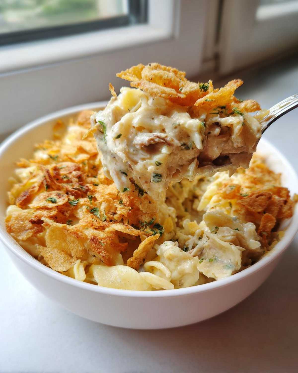 A spoonful of creamy Tuna Casserole, topped with crispy fried onions, being lifted from a white bowl.