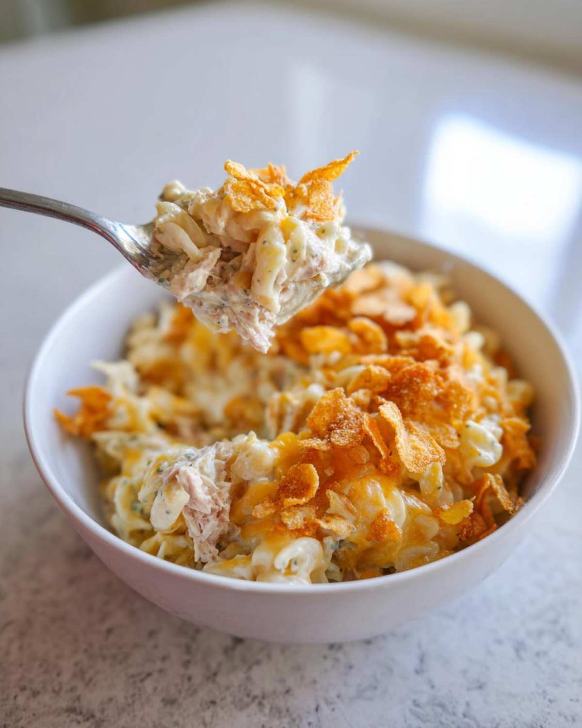A spoonful of creamy Tuna Casserole topped with crunchy cornflakes is lifted from a white bowl.