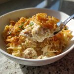 A spoonful of creamy Tuna Casserole, featuring egg noodles and a crunchy topping, being lifted from a white bowl.