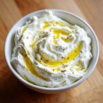 A close-up of creamy whipped feta dip topped with a generous swirl of golden olive oil and herbs.