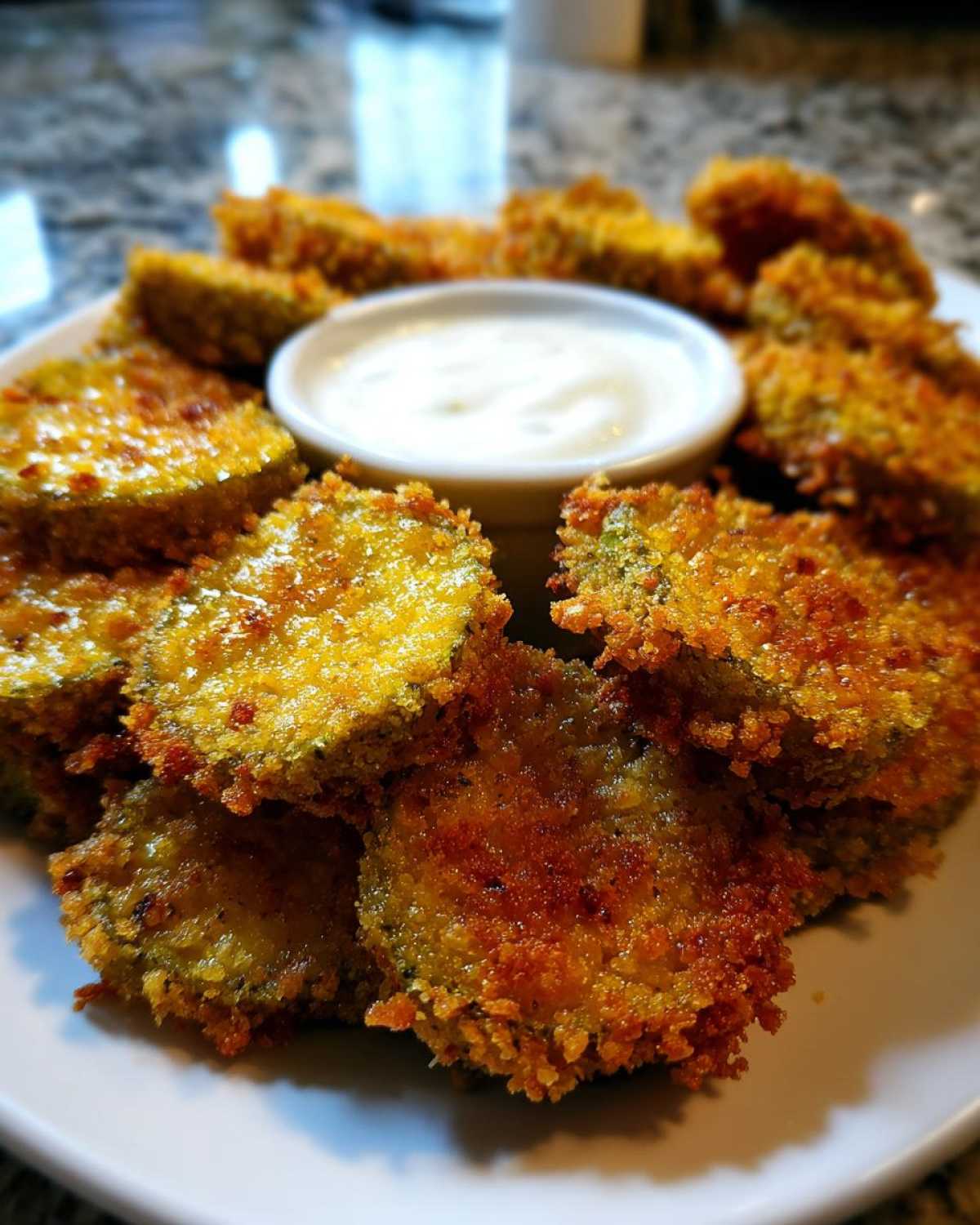 A plate piled high with golden brown, crispy Fried Pickles surrounding a small bowl of white dipping sauce.