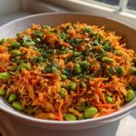 A vibrant bowl of orange-tinted crispy rice salad mixed with shredded carrots and bright green edamame, topped with fresh cilantro.
