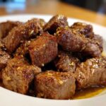 Close-up of tender, glazed cubes of crockpot steak served in a white bowl with garlic bits.