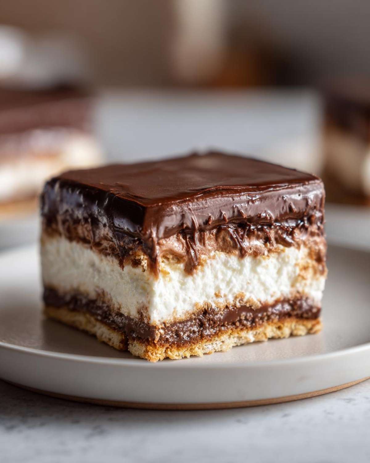Close-up of a square slice of eclair cake showing layers of crust, chocolate filling, white cream, and chocolate ganache topping.
