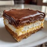 A square slice of eclair cake featuring layers of graham cracker, vanilla pudding, whipped cream, and chocolate frosting.