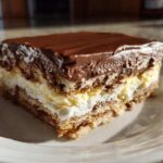 A square slice of layered eclair cake featuring graham crackers, vanilla cream, whipped topping, and chocolate ganache.
