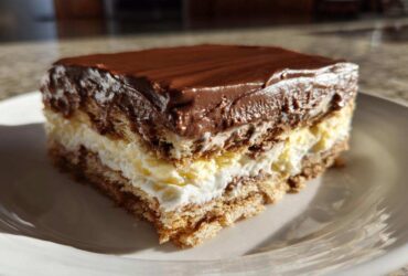 A square slice of layered eclair cake featuring graham crackers, vanilla cream, whipped topping, and chocolate ganache.