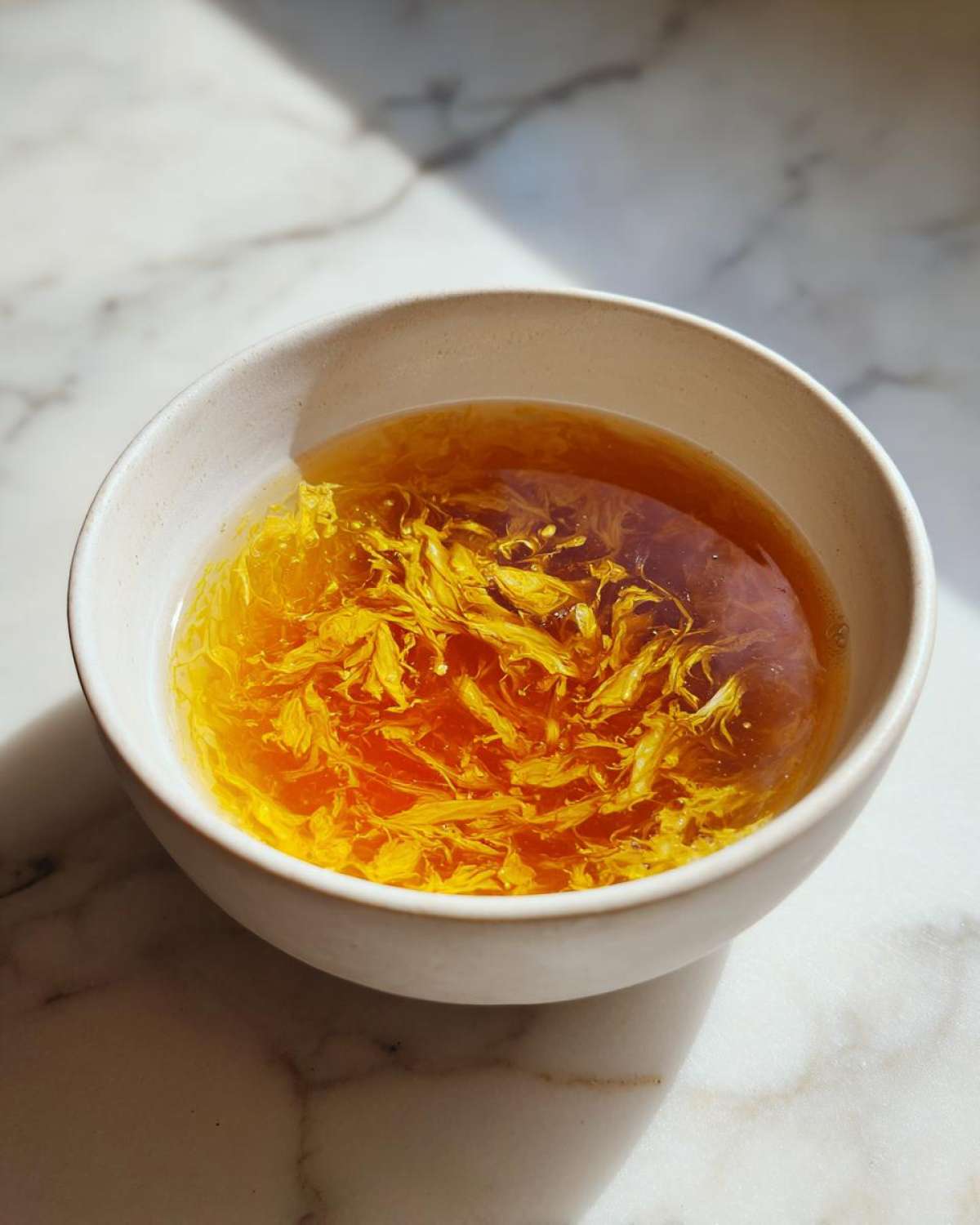 A close-up of a light-colored bowl filled with rich broth and delicate, bright yellow strands of egg drop soup.