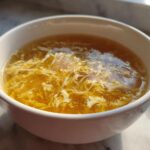 A close-up of a white bowl filled with golden broth and delicate ribbons of cooked egg, characteristic of egg drop soup.