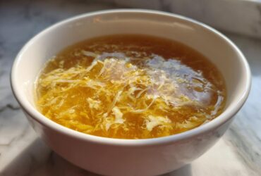 A close-up of a white bowl filled with golden broth and delicate ribbons of cooked egg, characteristic of egg drop soup.