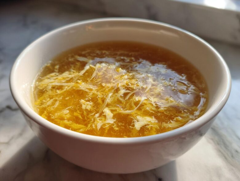 A close-up of a white bowl filled with golden broth and delicate ribbons of cooked egg, characteristic of egg drop soup.