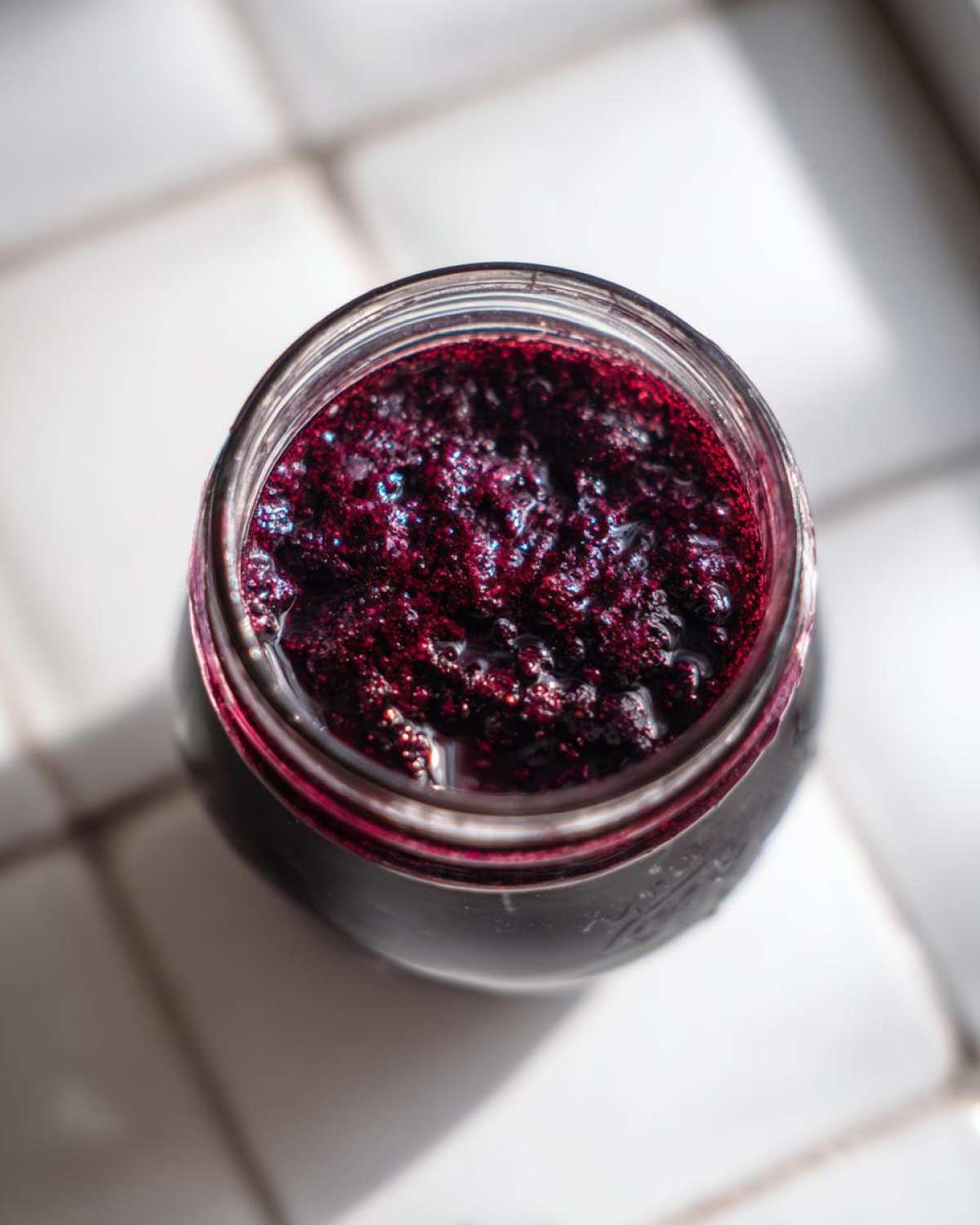 Overhead view of thick, dark purple elderberry syrup recipe filling a glass jar, resting on white tiles.