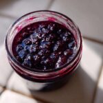 Overhead view of thick, dark purple elderberry syrup recipe mixture inside a clear glass jar, glistening in the sunlight.