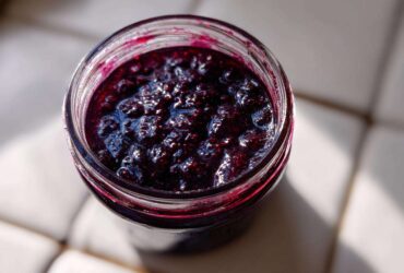 Overhead view of thick, dark purple elderberry syrup recipe mixture inside a clear glass jar, glistening in the sunlight.