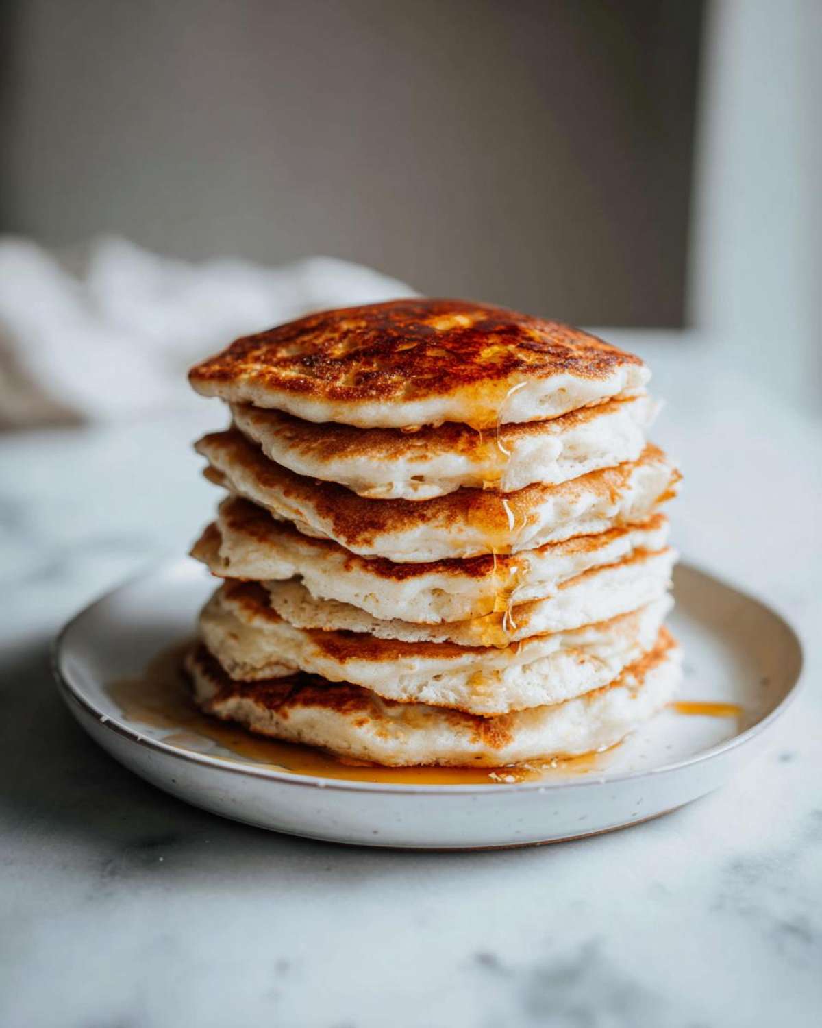 A tall stack of six fluffy oatmeal pancakes drizzled with golden syrup on a small white plate.