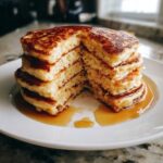 A tall stack of fluffy oatmeal pancakes cut open, drizzled generously with maple syrup on a white plate.