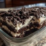 Close-up of a rich, layered slice of dirt cake featuring dark chocolate cake, white cream filling, and crushed cookie topping.
