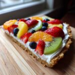 A close-up slice of fruit pizza sugar cookie topped with cream cheese frosting and fresh strawberries, kiwi, blueberries, and mandarin oranges.