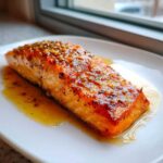 Close-up of a perfectly cooked, glazed air fryer salmon fillet resting in its sauce on a white plate.
