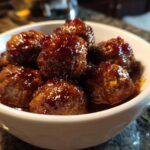 Close-up of shiny, glazed Grape Jelly Meatballs piled high in a small white serving bowl.