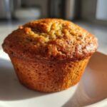 A single, golden-brown banana protein muffin sitting on a white dish, highlighted by bright sunlight.