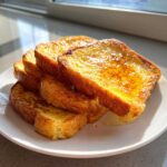 A stack of golden brown, syrup-drizzled french toast slices resting on a white plate in bright sunlight.