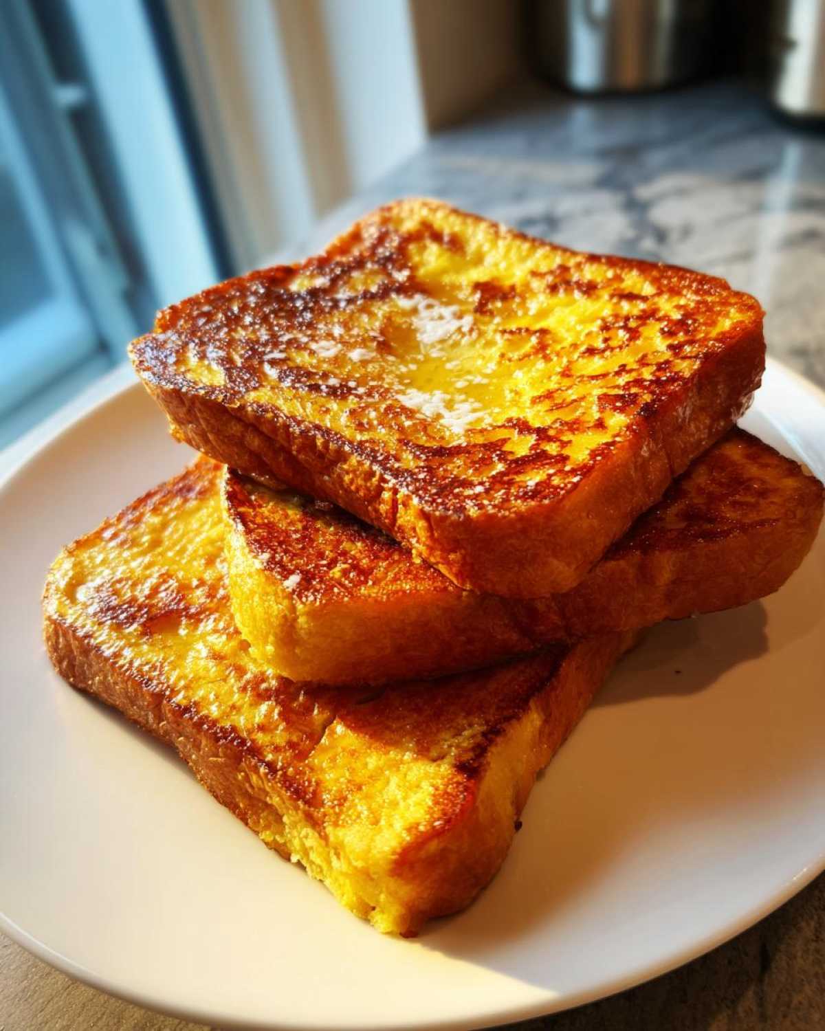 Three golden brown slices of perfectly cooked french toast stacked on a white plate, lightly dusted with powdered sugar.