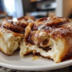 Two soft, gooey cinnamon rolls covered in caramel glaze and white frosting, one pulled apart to show the filling.