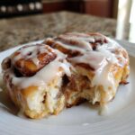 A close-up shot of a freshly baked cinnamon roll, covered in white icing, with a bite taken out showing the soft interior.