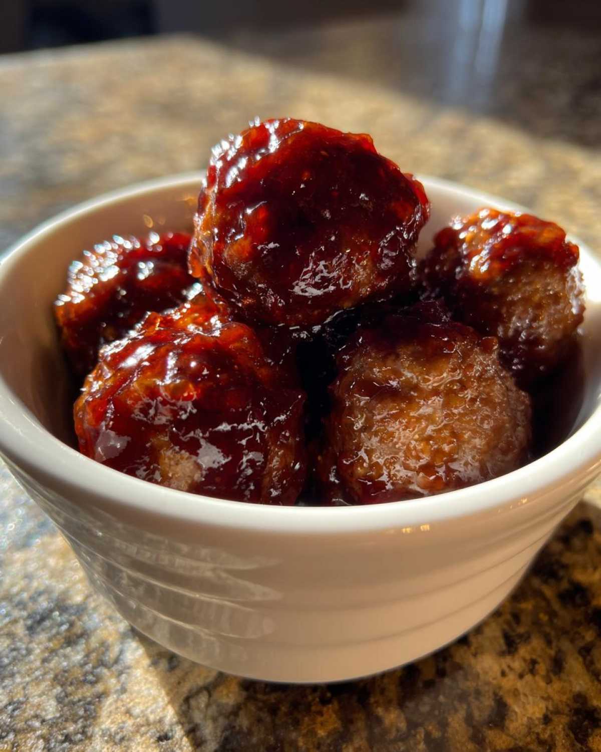 A small white bowl filled with shiny, dark red Grape Jelly Meatballs, glistening under direct light.