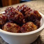 Close-up of several savory grape jelly meatballs coated in a thick, glossy, dark red sauce served in a white bowl.