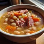 Close-up of a steaming bowl of Ham and bean soup featuring white beans, chunks of ham, carrots, and celery.