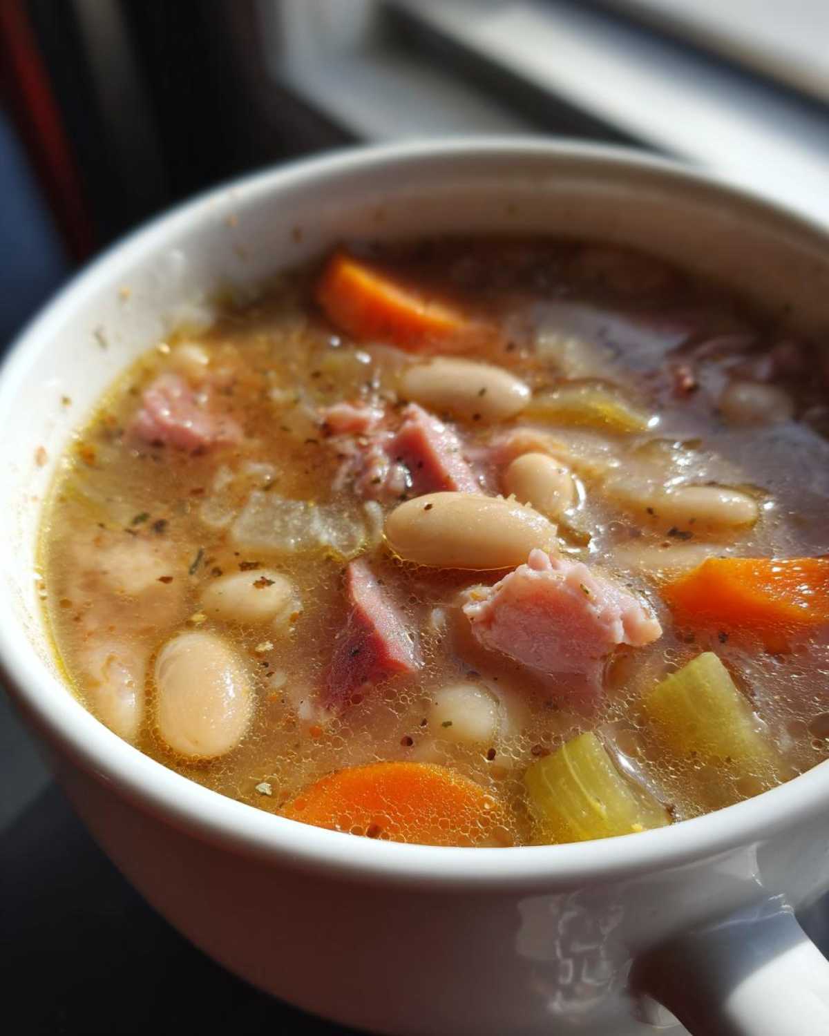 Close-up view of a white bowl filled with steaming Ham and bean soup, featuring white beans, chunks of ham, carrots, and celery.