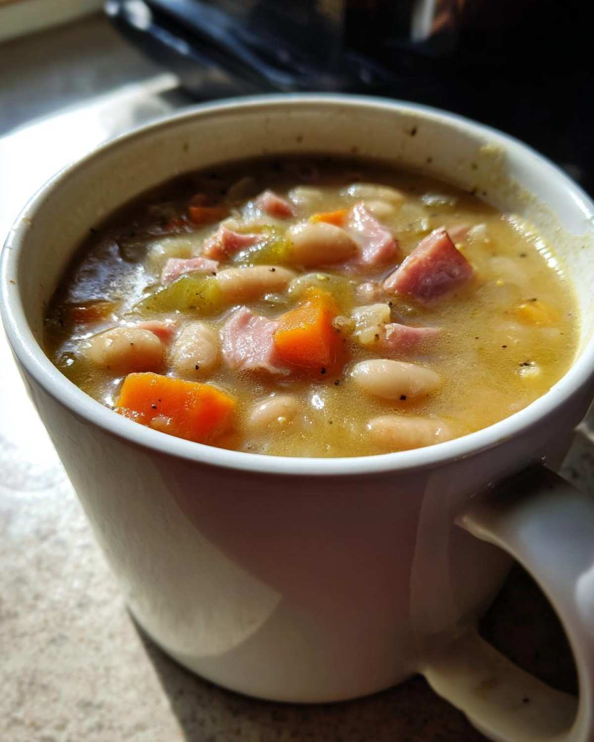 Close-up of hearty Ham and bean soup filled with white beans, diced ham, and carrots in a white mug.