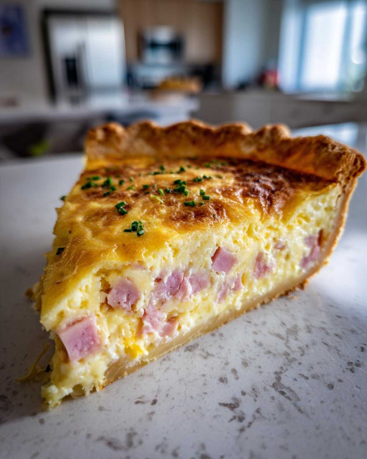 A close-up of a thick slice of golden-brown ham and cheese quiche, garnished with fresh chives.