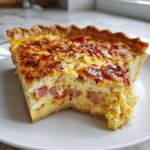 A close-up of a thick slice of ham and cheese quiche showing the flaky crust and savory egg filling.