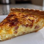 A close-up of a golden-brown slice of ham and cheese quiche on a white plate.