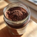 Close-up of rich brown homemade hot cocoa mix powder filling a clear glass jar.