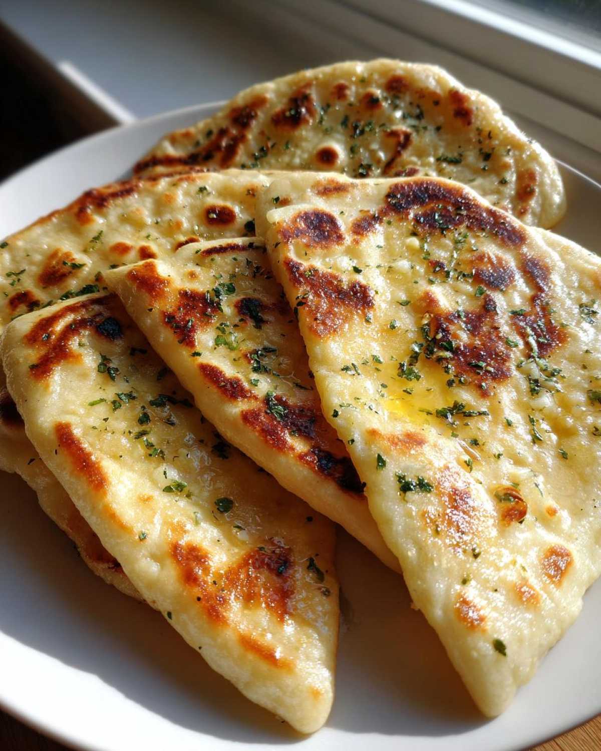 A stack of freshly cooked homemade naan bread pieces, brushed with melted butter and sprinkled with green herbs.