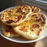 A stack of four golden, puffy homemade naan bread pieces brushed with melted butter and herbs.