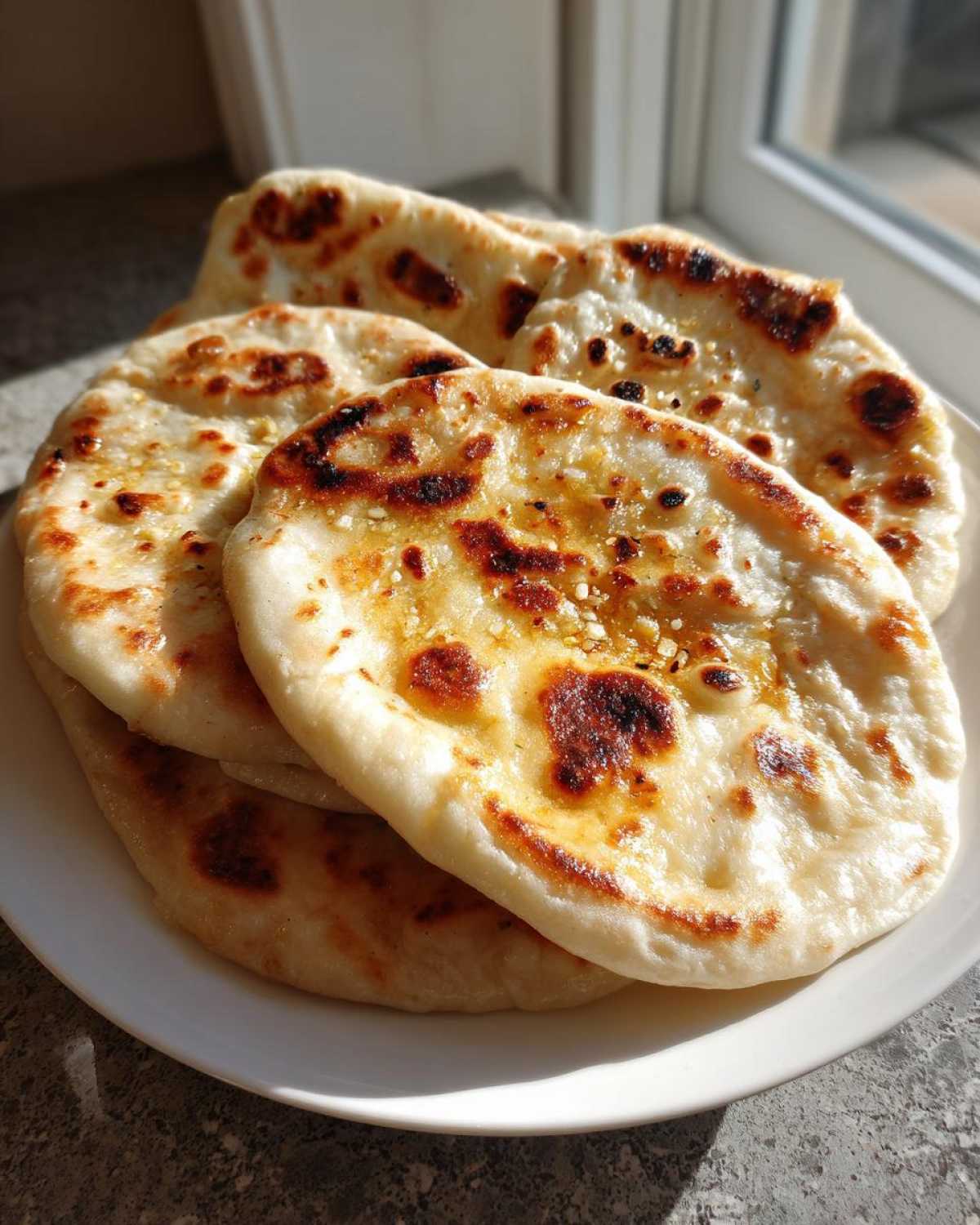 A stack of fluffy, golden-brown homemade naan bread brushed with butter, resting on a white plate.